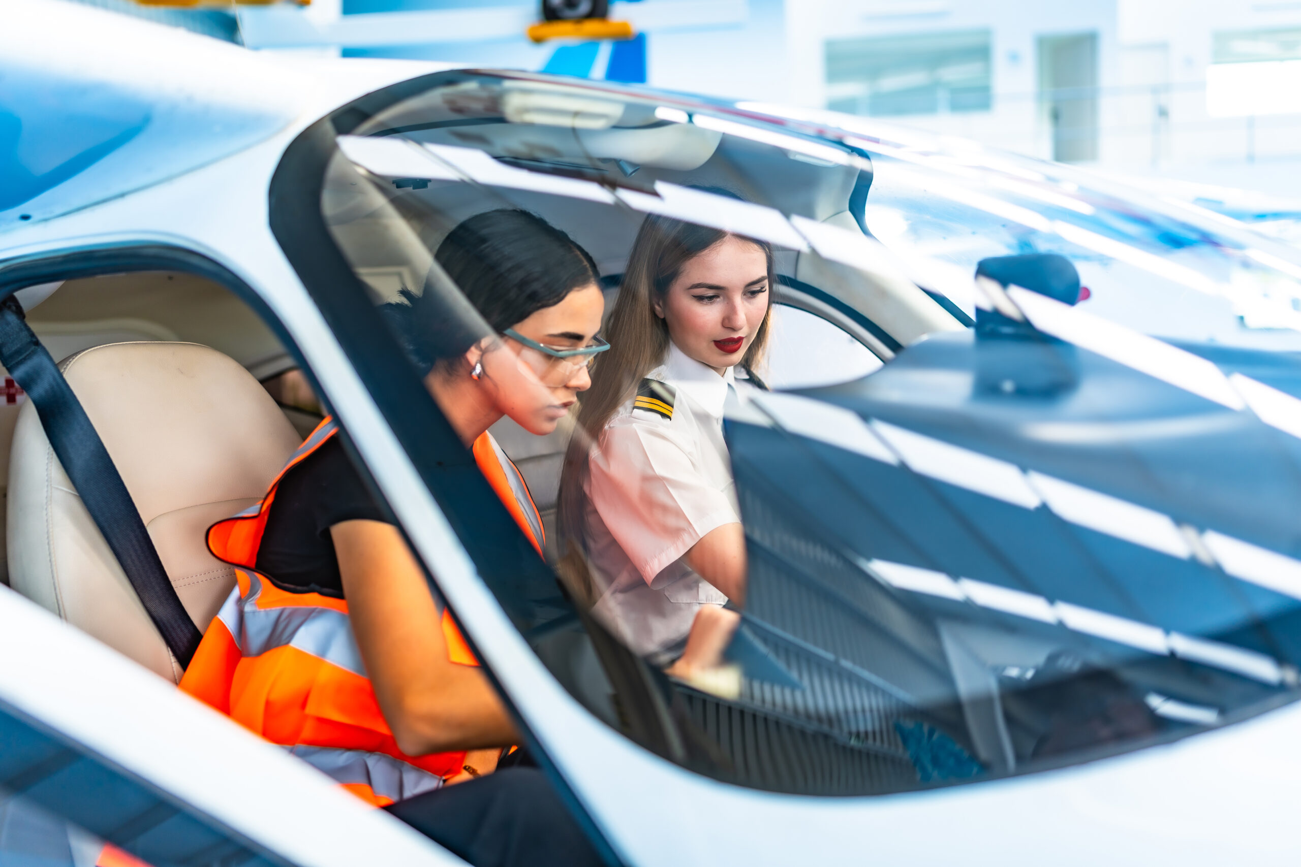 Two female pilots, one in uniform and the other in a safety vest, collaborate inside an aircraft cockpit, focusing on flight controls