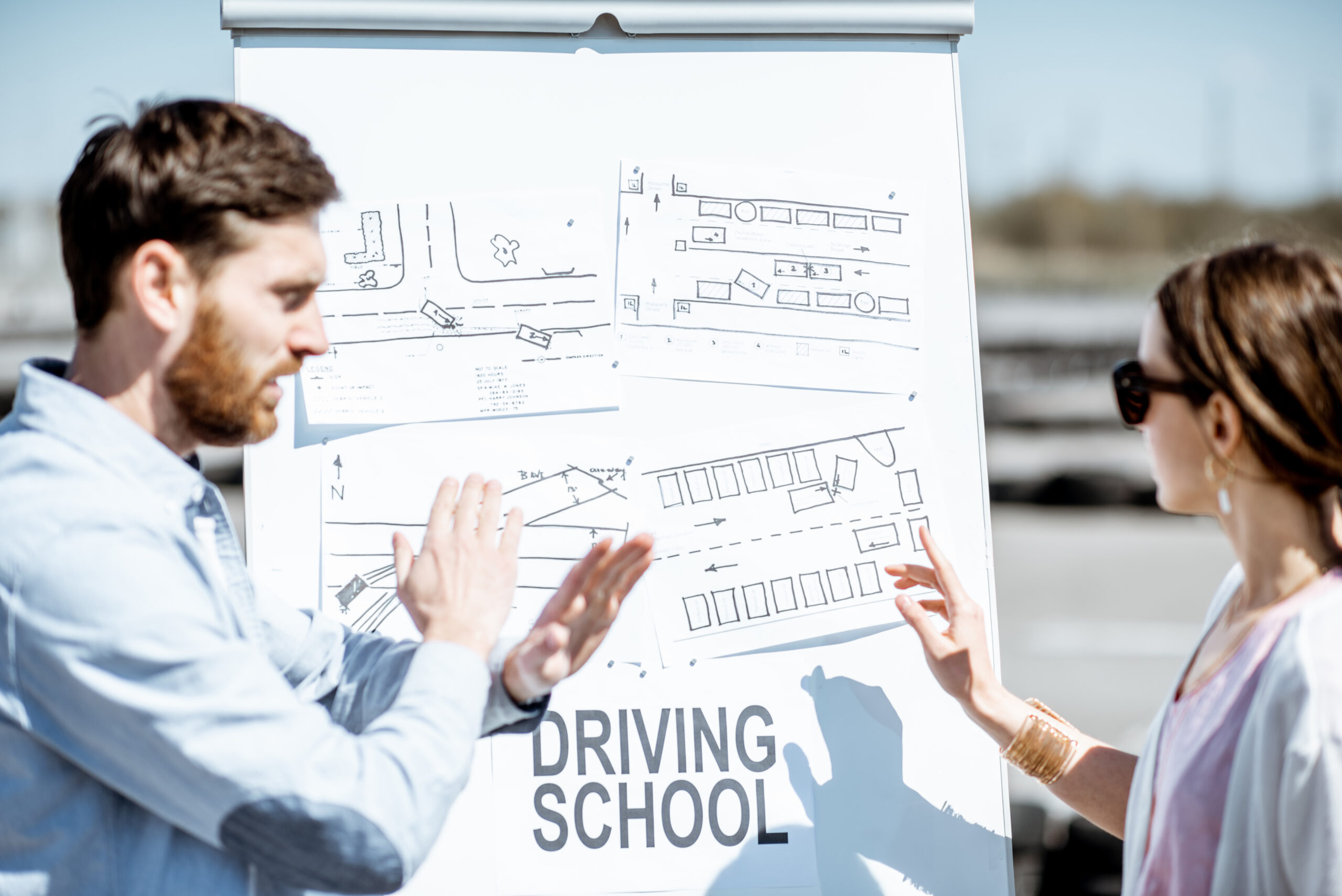 Male instructor showing traffic shemes to a young female student standing on the training ground at the driver's school
